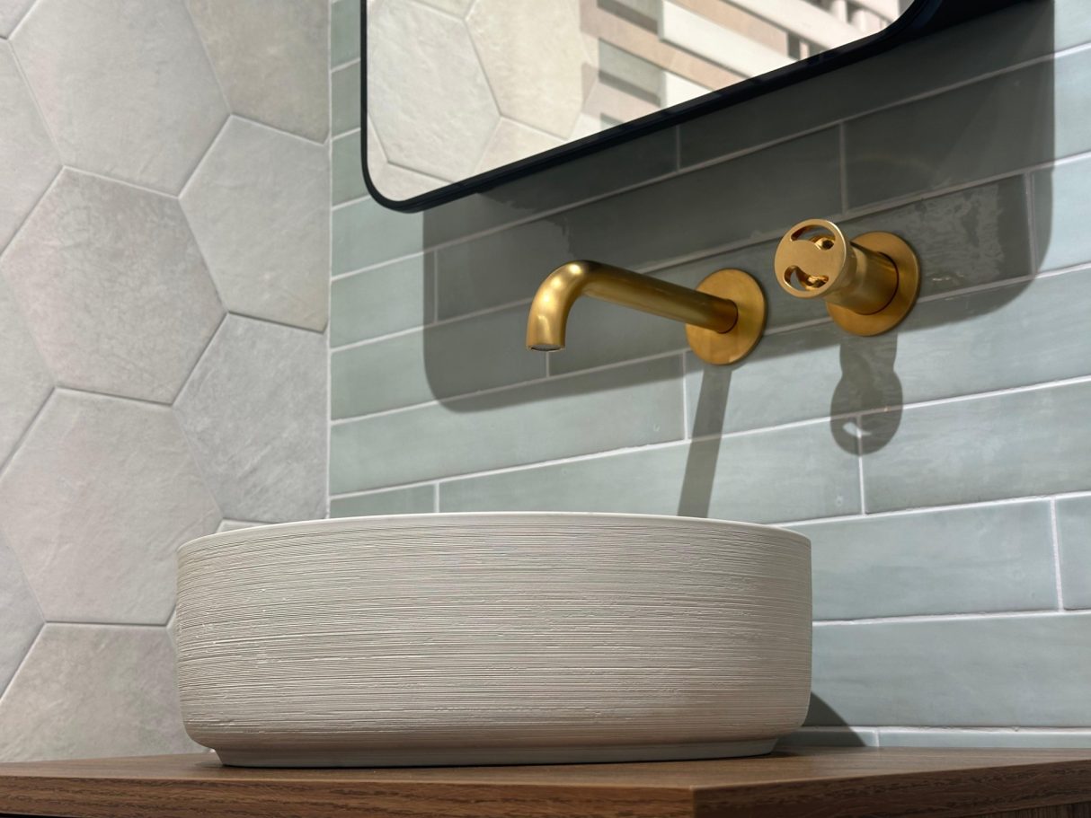 Lavabo moderno de cerámica con grifería dorada y espejo en una pared de azulejos hexagonales.